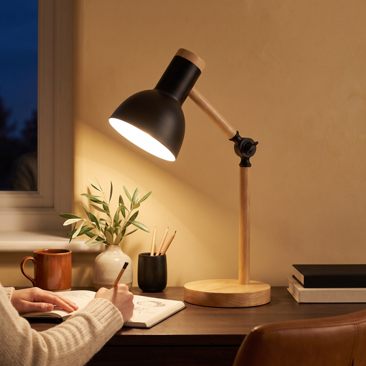 Ingrid wood desk lamp in black with adjustable oak arm, lit on a walnut desk at night